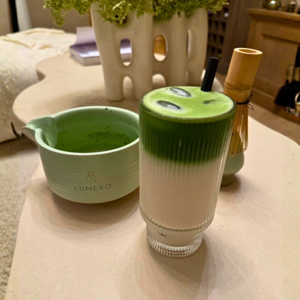 Matcha latte in glazen mok met schuimlaag op tafel, naast matcha kom en accessoires.