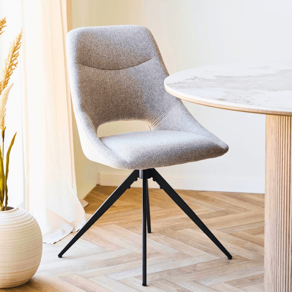 House Nordic Dokka eetkamerstoel in zandkleurige stof met zwarte poten, geplaatst aan een ronde eettafel in Scandinavisch interieur met houten vloer en lichte gordijnen.