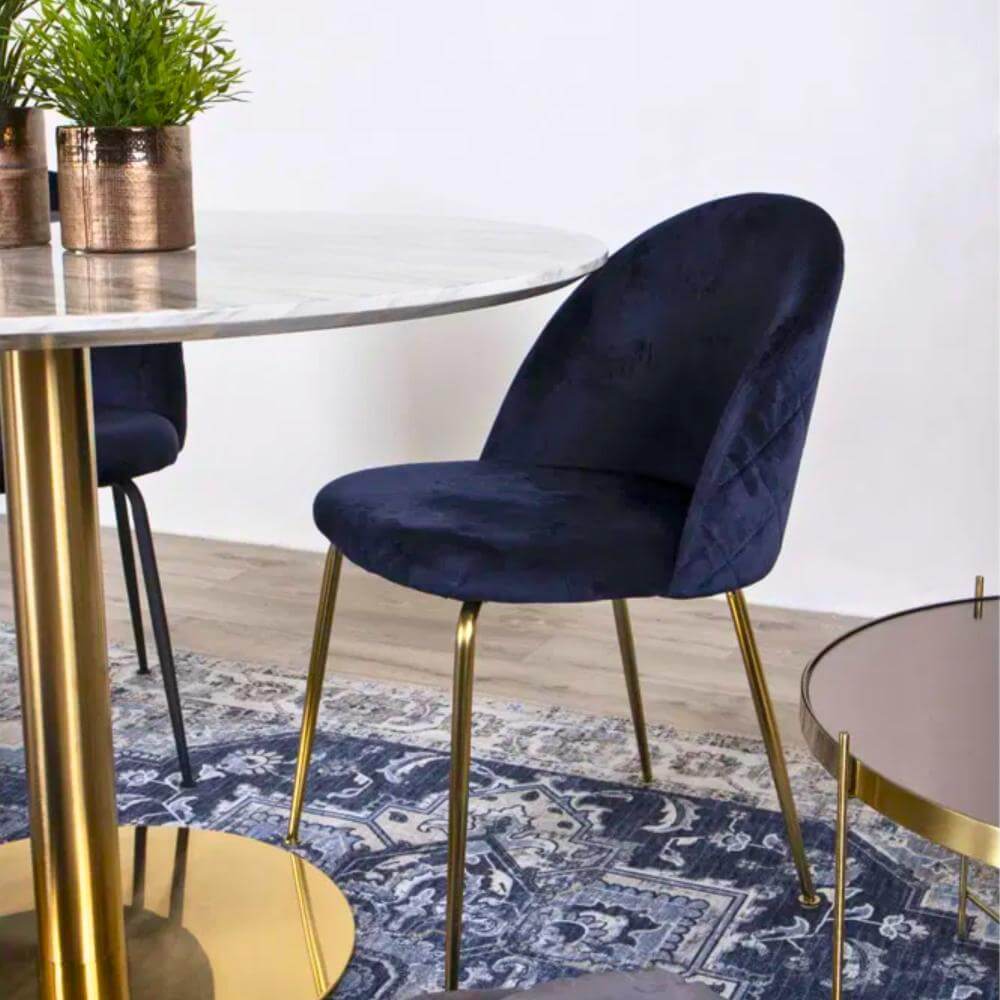 Close-up van de donkerblauwe velvet Geneve eetkamerstoel van House Nordic met gouden poten aan een ronde marmeren eettafel – stijlvolle Scandinavische interieurfoto van Lunero.