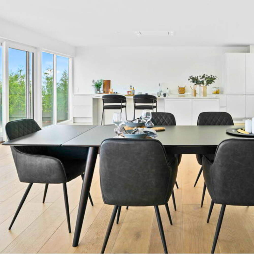 Scandinavische eetkamer met House Nordic Harbo eetkamerstoelen in donkergrijs PU-leer en zwarte poten, geplaatst rond een moderne eettafel in een lichte keuken met houten vloer.