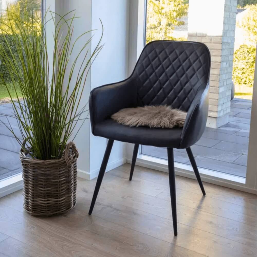 Zwarte House Nordic Harbo eetkamerstoel van PU-leer met ruitpatroon en zacht vachtkussen, geplaatst naast een rotan plantenmand in een modern Scandinavisch interieur.