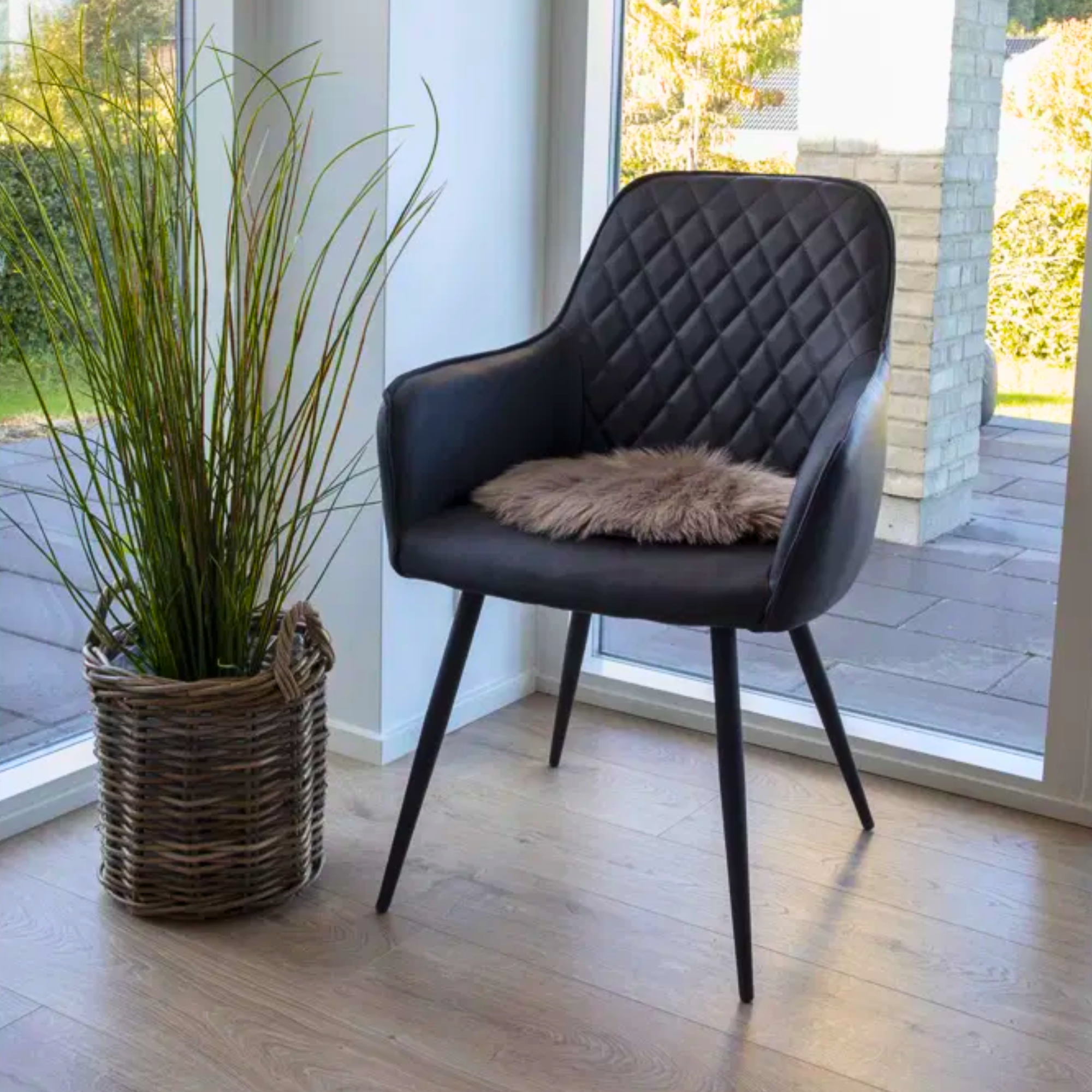 Zwarte House Nordic Harbo eetkamerstoel van PU-leer met ruitpatroon en zacht vachtkussen, geplaatst naast een rotan plantenmand in een modern Scandinavisch interieur.