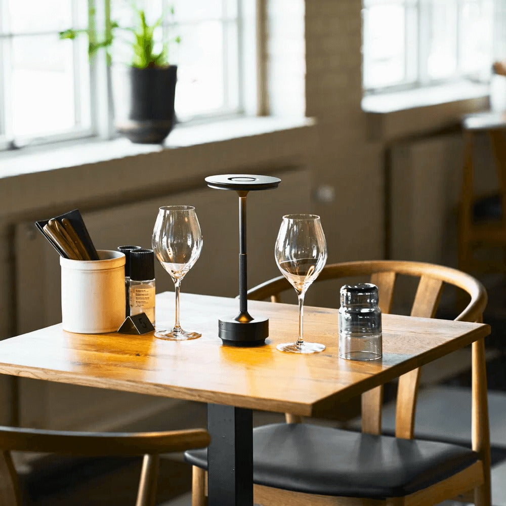House Nordic Lyneham LED tafellamp in zwart op een gedekte restauranttafel met glazen en houten stoelen in een sfeervolle horecazaak.