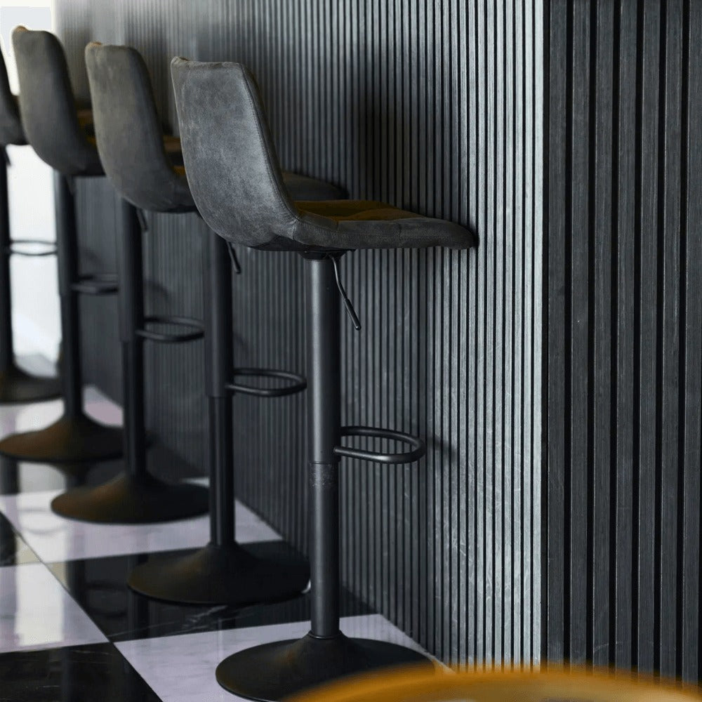Donkergrijze House Nordic Middelfart barkrukken met zwarte metalen poten, geplaatst aan moderne zwarte barwand met verticale panelen.