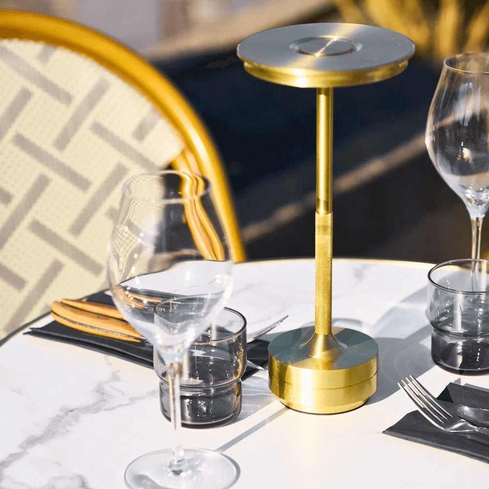 Gouden Lyneham LED tafellamp op een gedekte marmeren terrastafel in horecasetting, tussen glazen en bestek.
