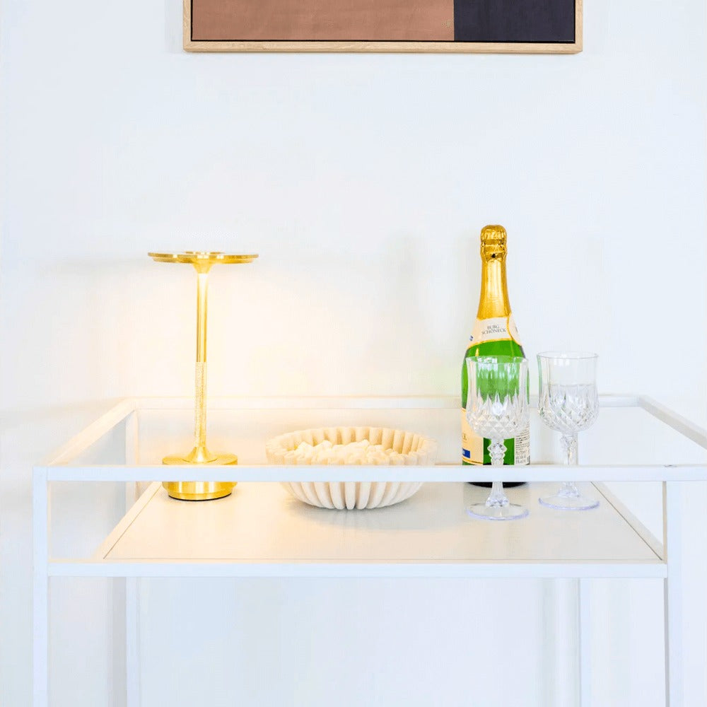 Gouden Lyneham LED tafellamp van House Nordic op een witte bartafel, gestyled met champagne en kristallen glazen.