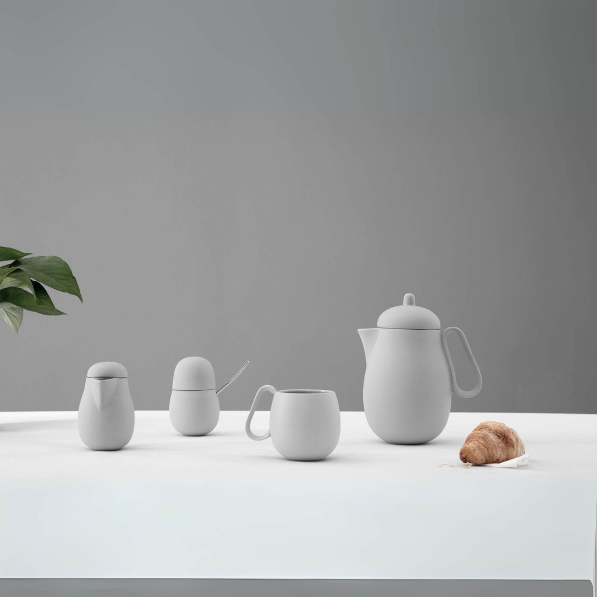 Minimalistische witte keramische theeset met theepot, melkkan, suikerpot en kopje op tafel met croissant.