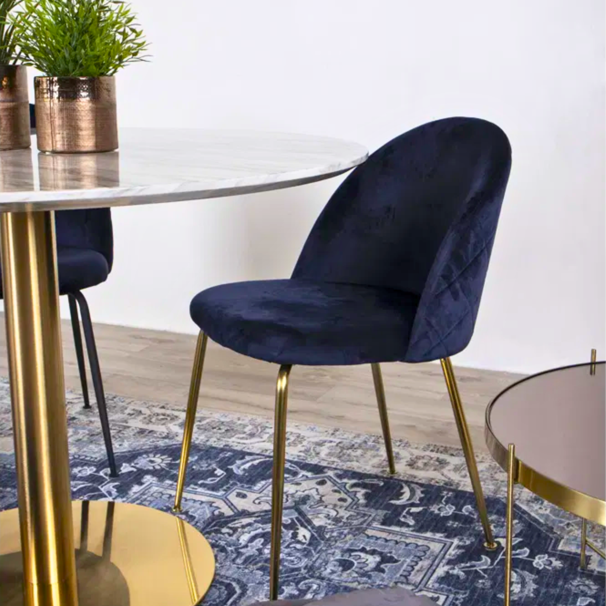Close-up van de donkerblauwe velvet Geneve eetkamerstoel van House Nordic met gouden poten aan een ronde marmeren eettafel – stijlvolle Scandinavische interieurfoto van Lunero.
