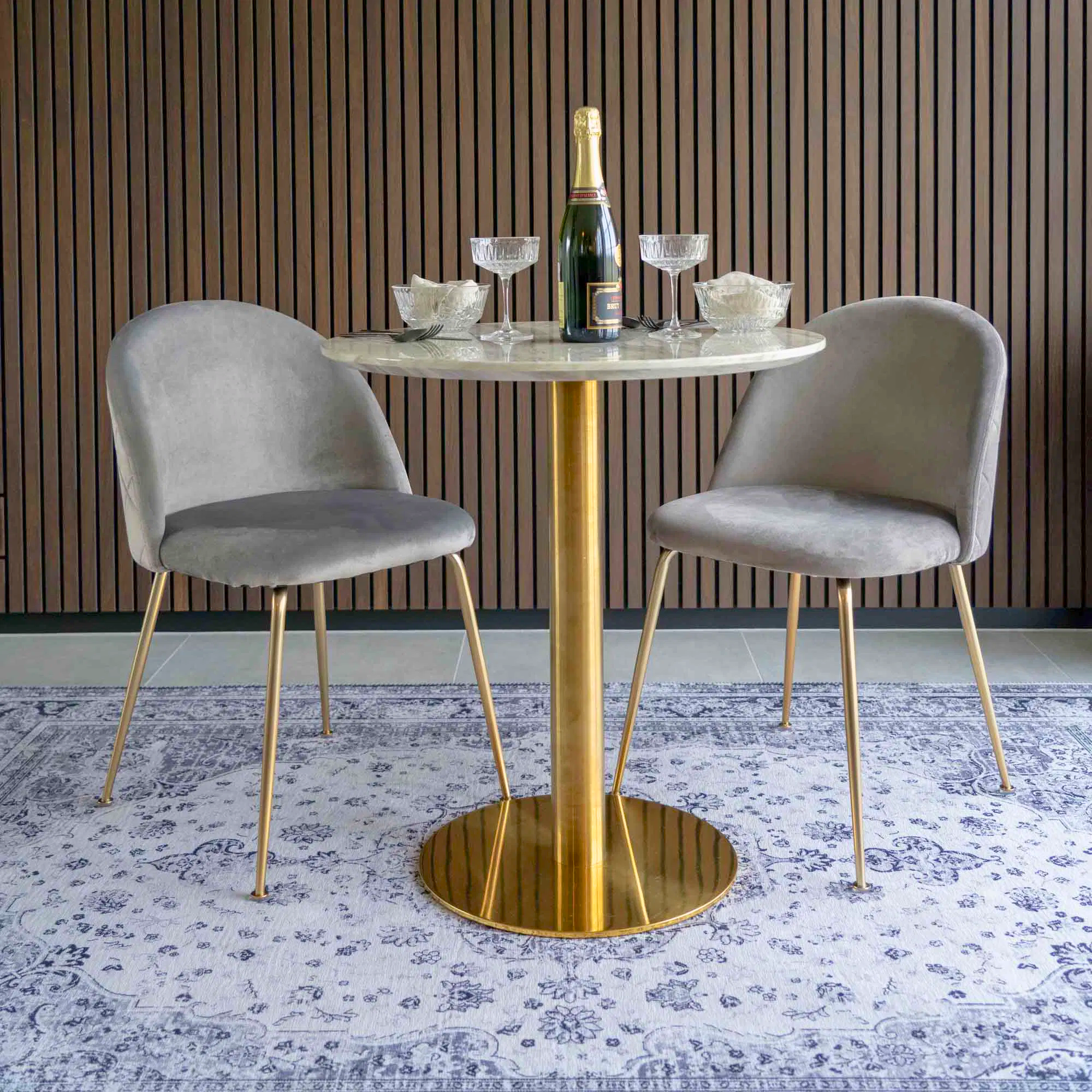 Twee grijs velvet House Nordic Geneve eetkamerstoelen met gouden poten aan een marmeren eettafel in modern interieur met houten wand.