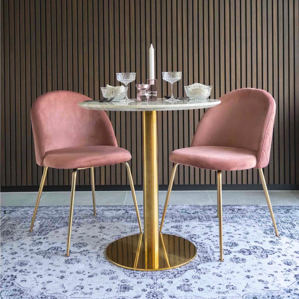 Twee roze velvet House Nordic Geneve eetkamerstoelen met gouden poten aan een marmeren eettafel in modern interieur met houten wand.