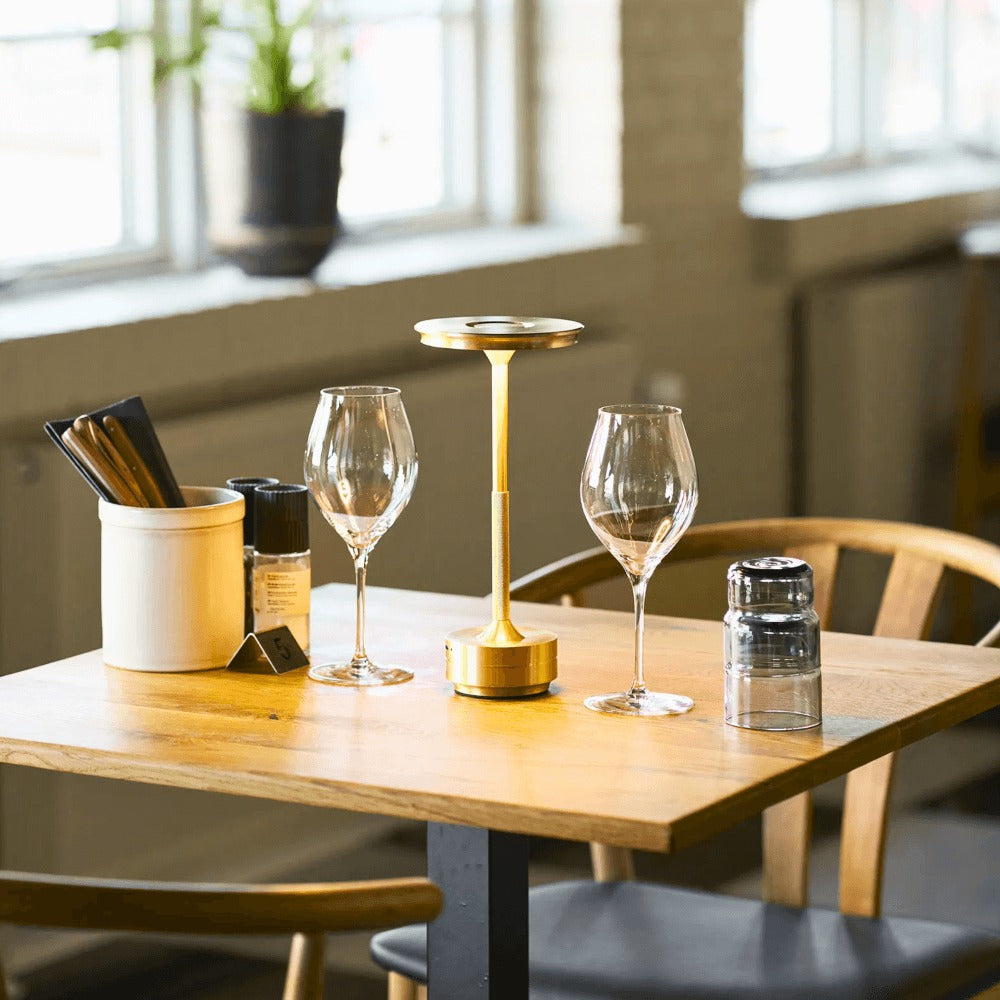 House Nordic Lyneham LED tafellamp in goud op een gedekte restauranttafel met glazen en houten stoelen in een sfeervolle horecazaak.