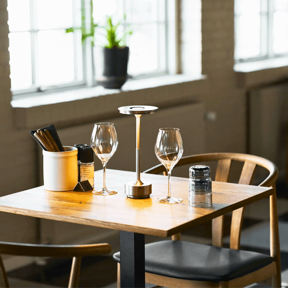 House Nordic Lyneham LED tafellamp in zilver op een gedekte restauranttafel met glazen en houten stoelen in een sfeervolle horecazaak.