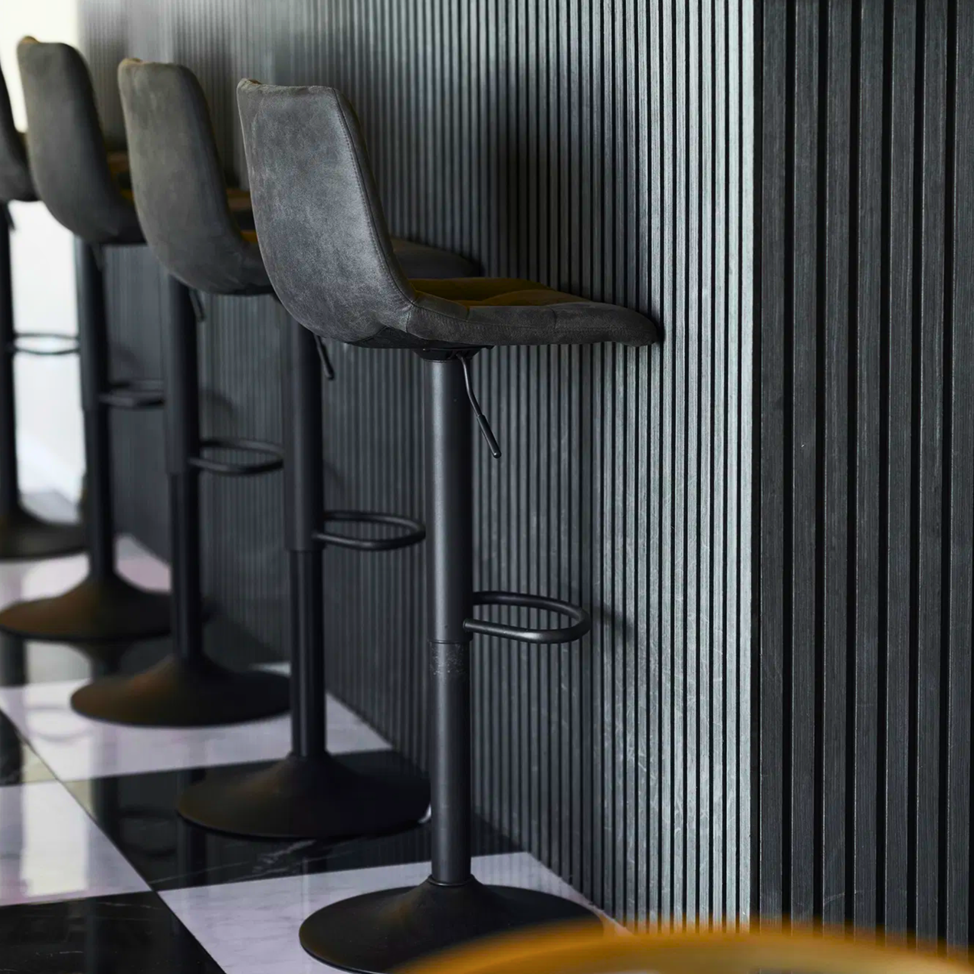 Donkergrijze House Nordic Middelfart barkrukken met zwarte metalen poten, geplaatst aan moderne zwarte barwand met verticale panelen.