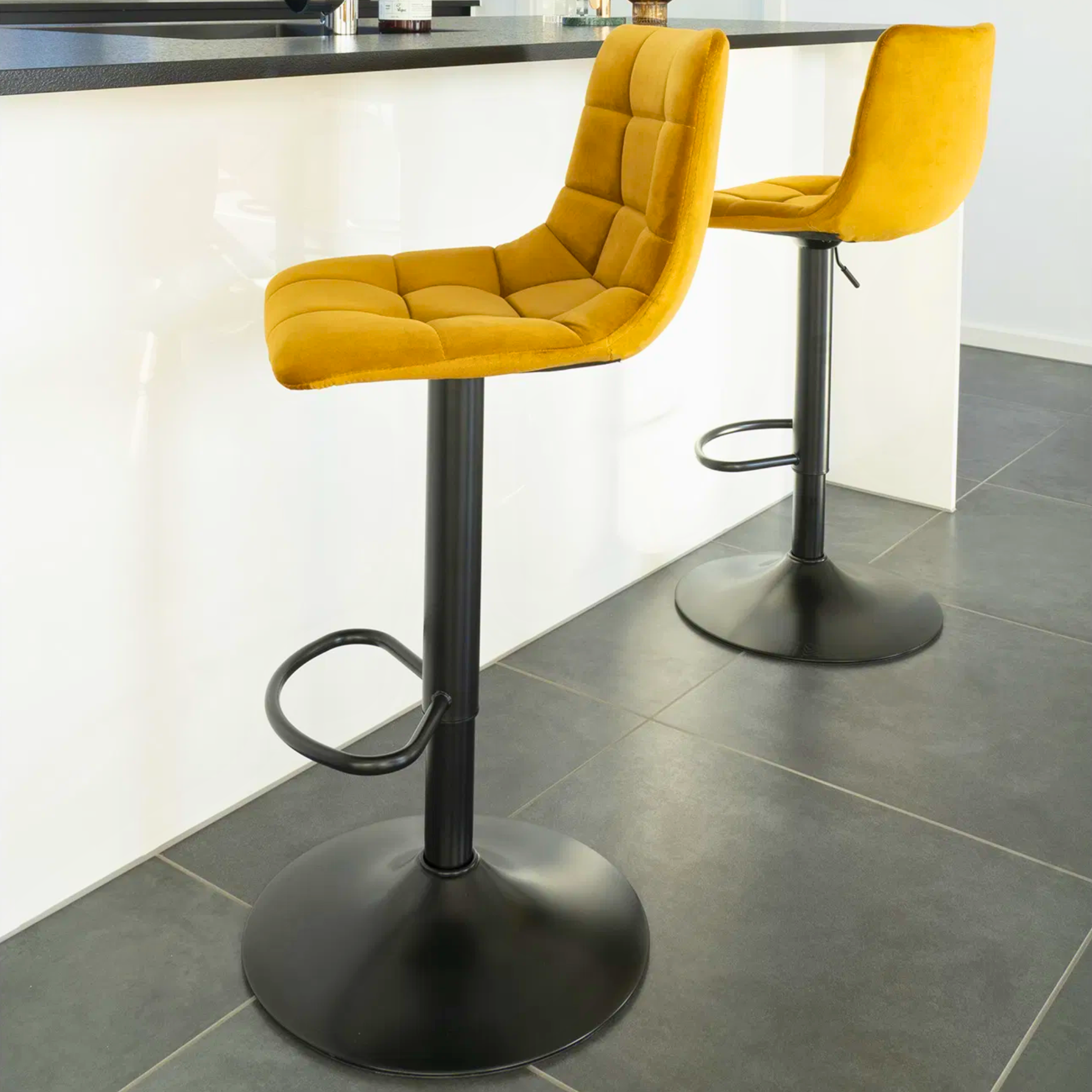 Twee House Nordic Middelfart barkrukken in geel velvet met zwarte metalen poten, geplaatst aan moderne witte keukenbar met grijze tegelvloer.