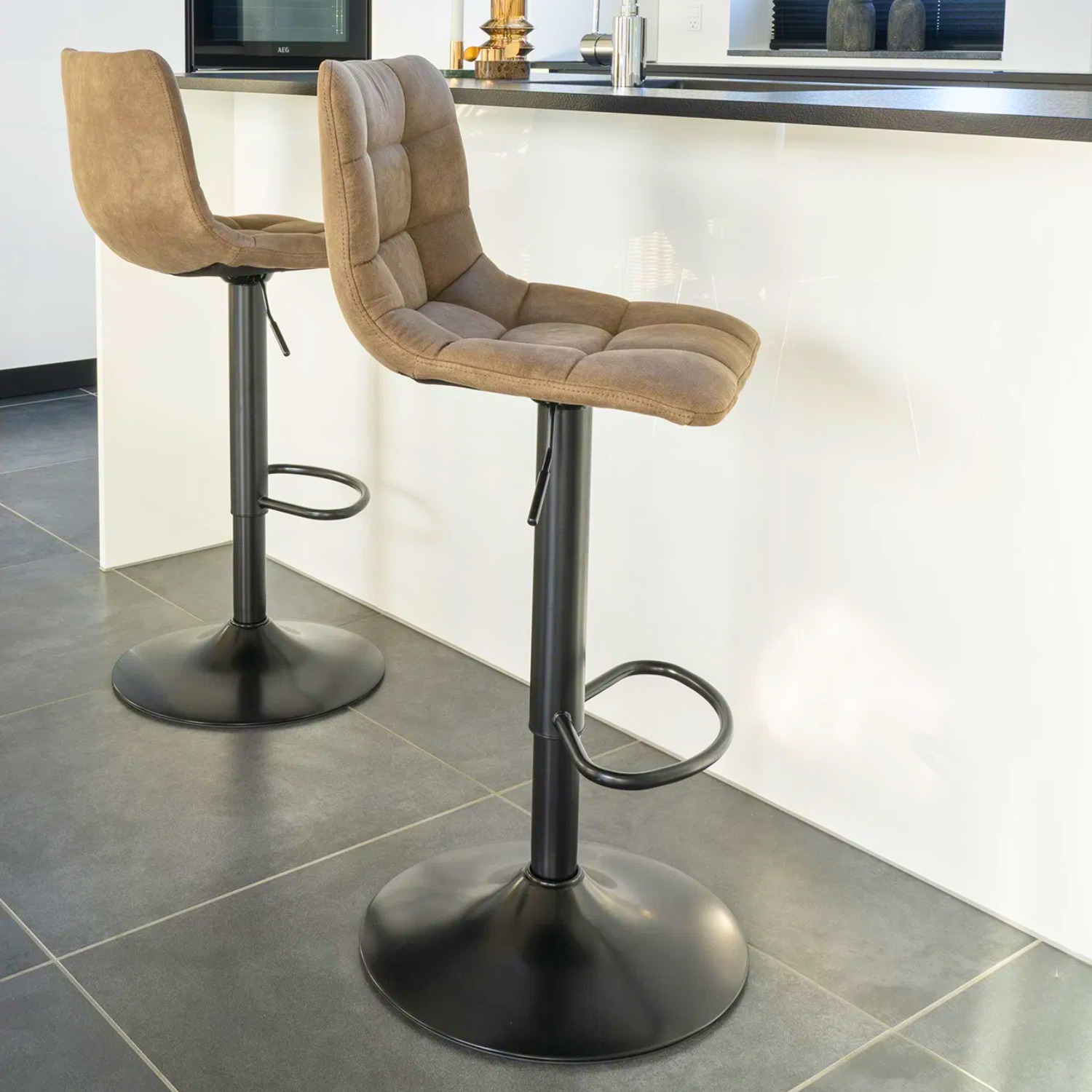 Twee House Nordic Middelfart barkrukken in lichtbruin microvezel met zwarte metalen poten, geplaatst aan moderne witte keukenbar met grijze tegelvloer.