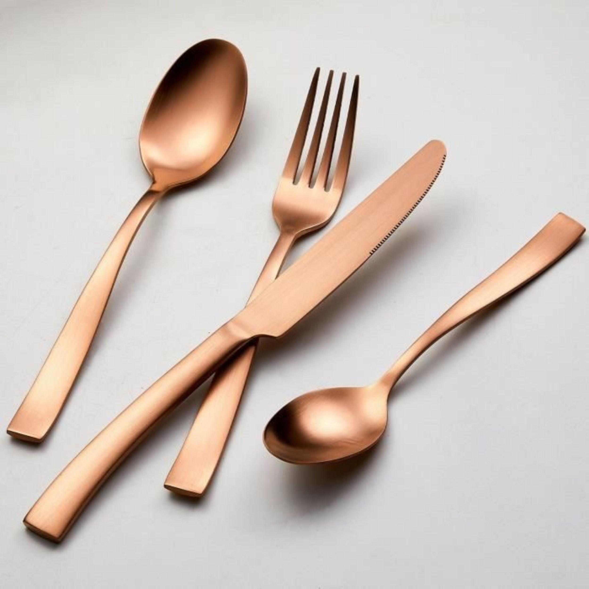 Koperkleurige RVS bestekset van Arthur Martin – flatlay foto met mes, vork, eetlepel en theelepel op lichtgrijze achtergrond.