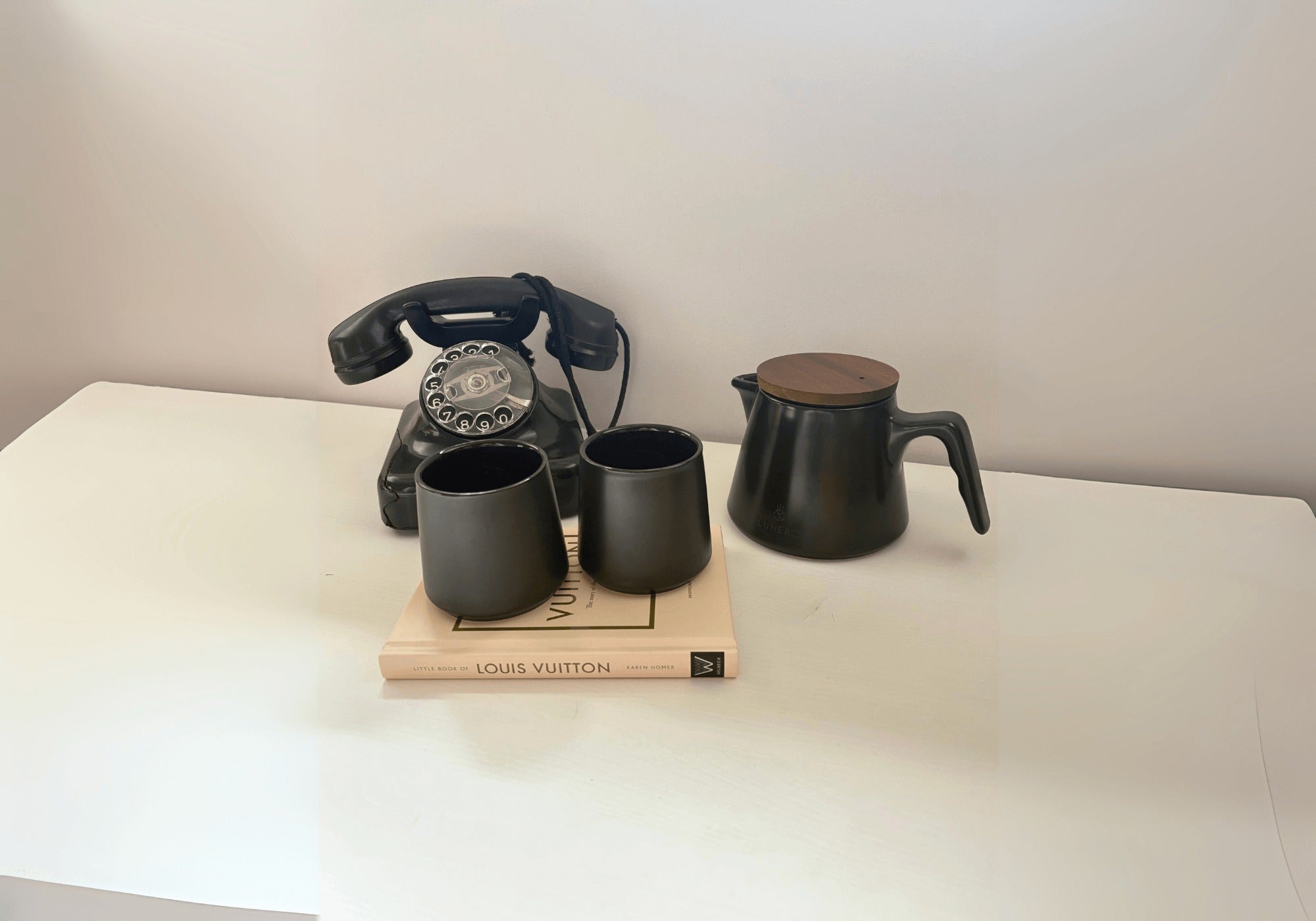 Lunero zwarte theeset met houten deksel en twee kopjes, naast retro telefoon en Louis Vuitton boek – stijlvolle contactpagina sfeerfoto.