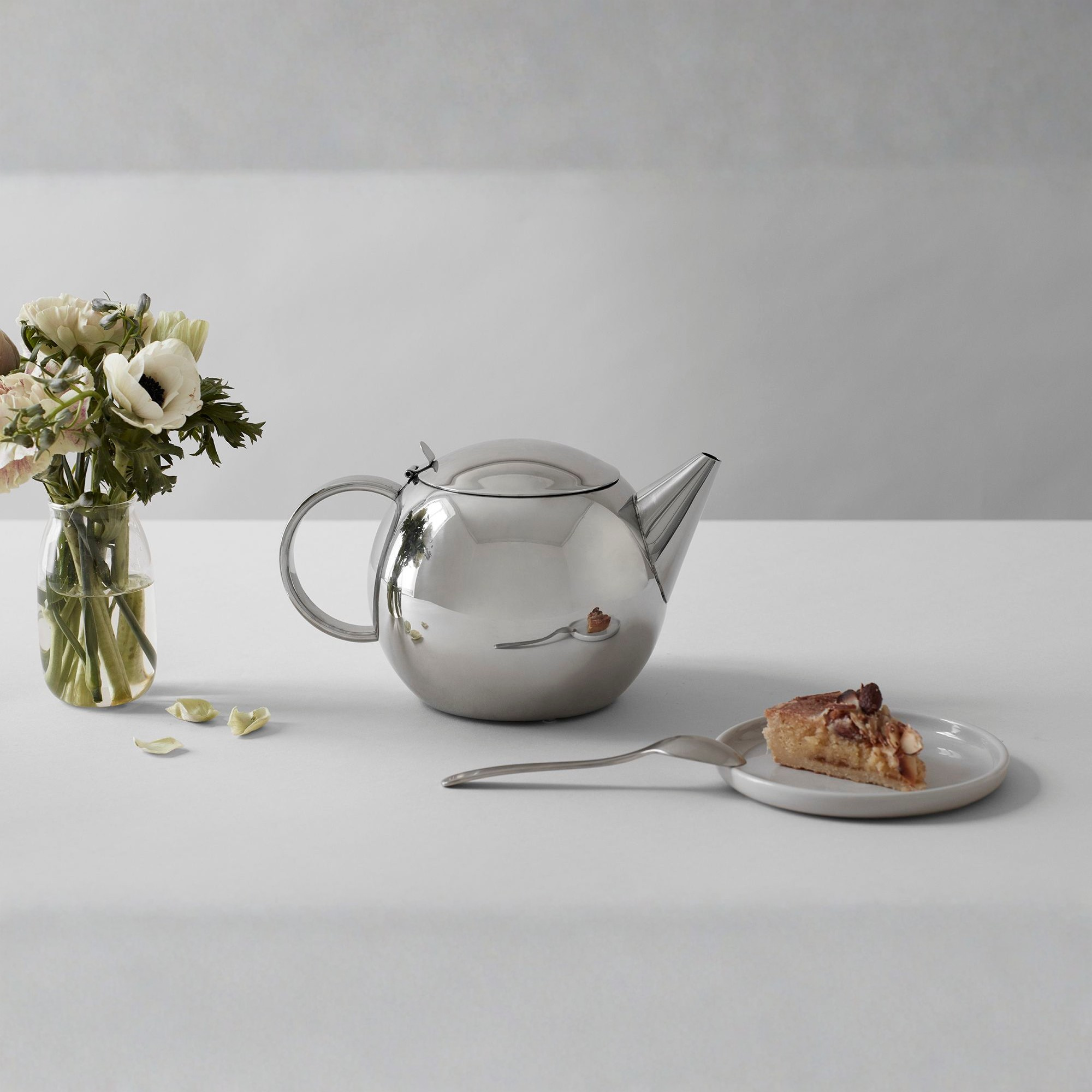 Viva Scandinavia Lucas theepot van roestvrij staal op tafel naast bloemenvaas en stuk taart – Scandinavisch design.